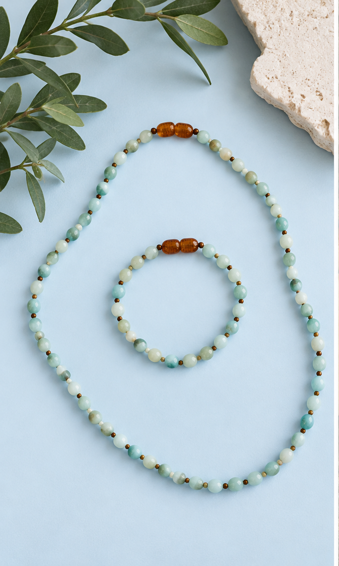 Acacia Amazonite necklace and matching bracelet with small blue-green beads and amber-toned accents arranged on a pale blue background, The Riann Ashlyn Collection.
