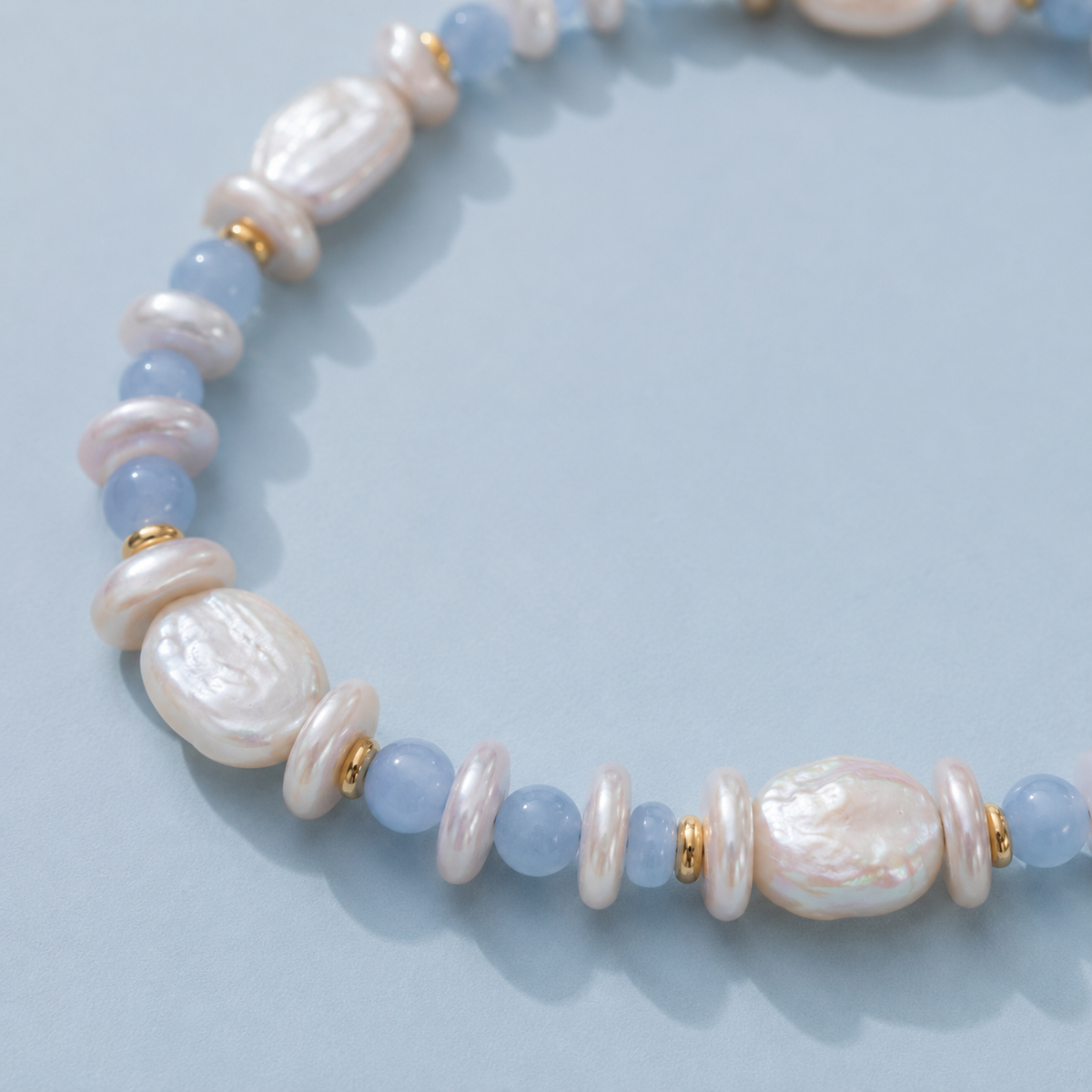 Close-up of white baroque pearls, light blue round beads and gold-tone spacers on a light blue background