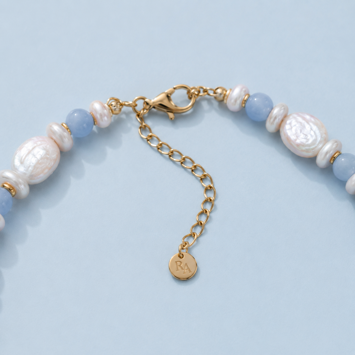 Close-up of light blue bead and white pearl necklace with gold-tone extender chain and round tag on light blue background