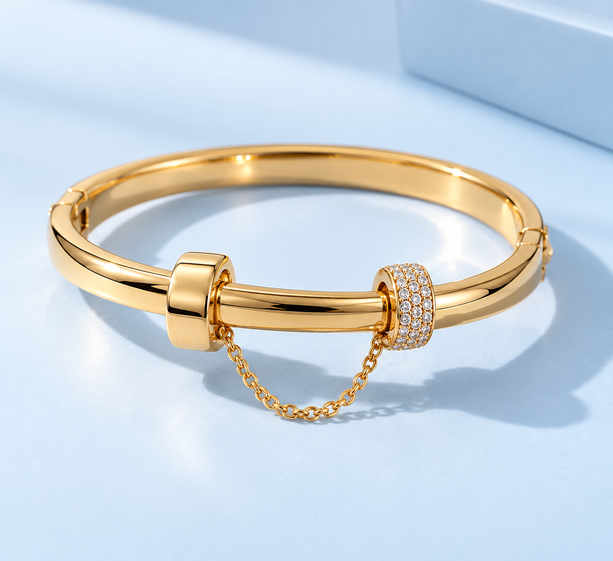 Channing Sculptural Bangle Bracelet from The Riann Ashlyn Collection, shown in a gold-tone finish with pavé accents and a connecting chain against a soft blue background.