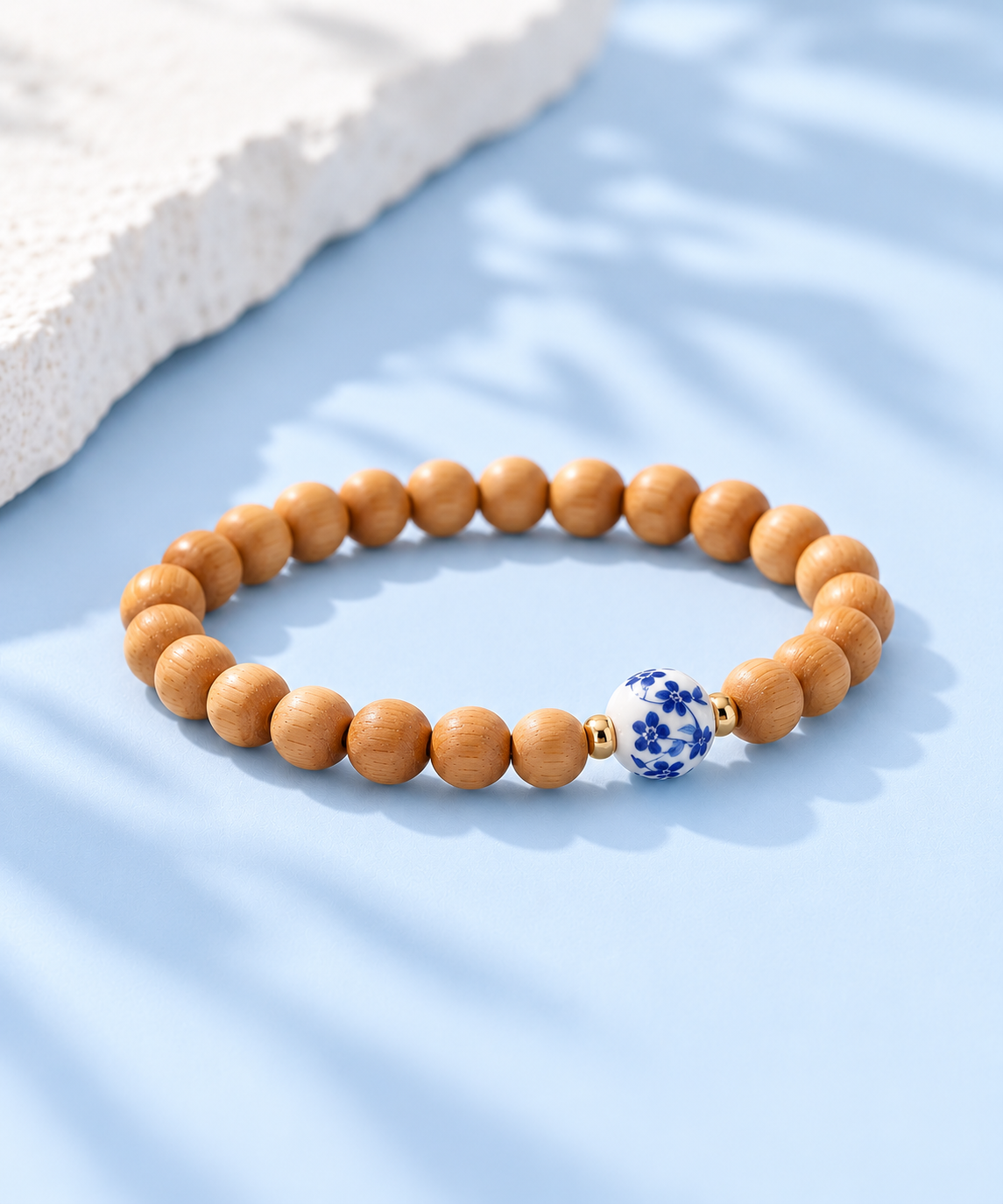 Angled view of Cypress cypress wood bracelet with blue floral porcelain centerpiece and gold-filled beads on pale blue background, The Riann Ashlyn Collection.