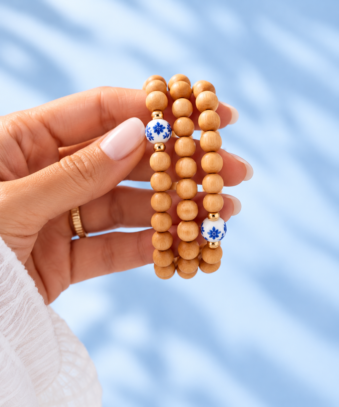 Hand holding three Cypress wood bead bracelets with blue-and-white floral porcelain accents and 14K gold-filled beads against a pale blue background, The Riann Ashlyn Collection.