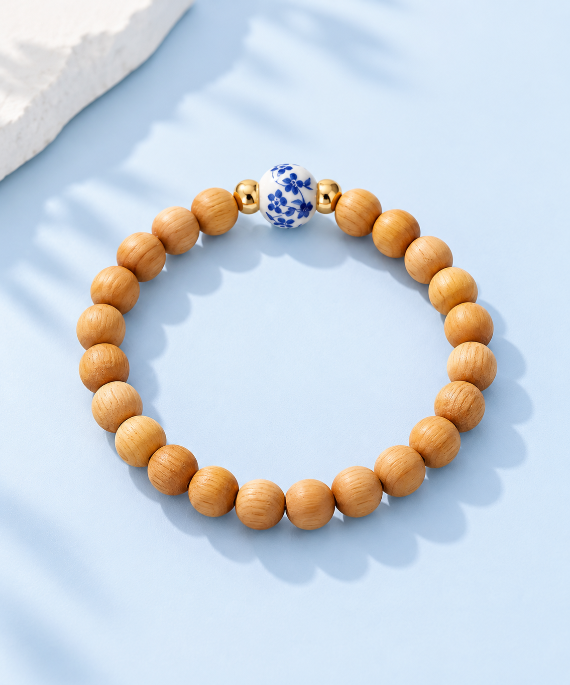 Cypress wood bead bracelet with hand-painted blue floral porcelain bead and 14K gold-filled accents on a pale blue background, The Riann Ashlyn Collection.