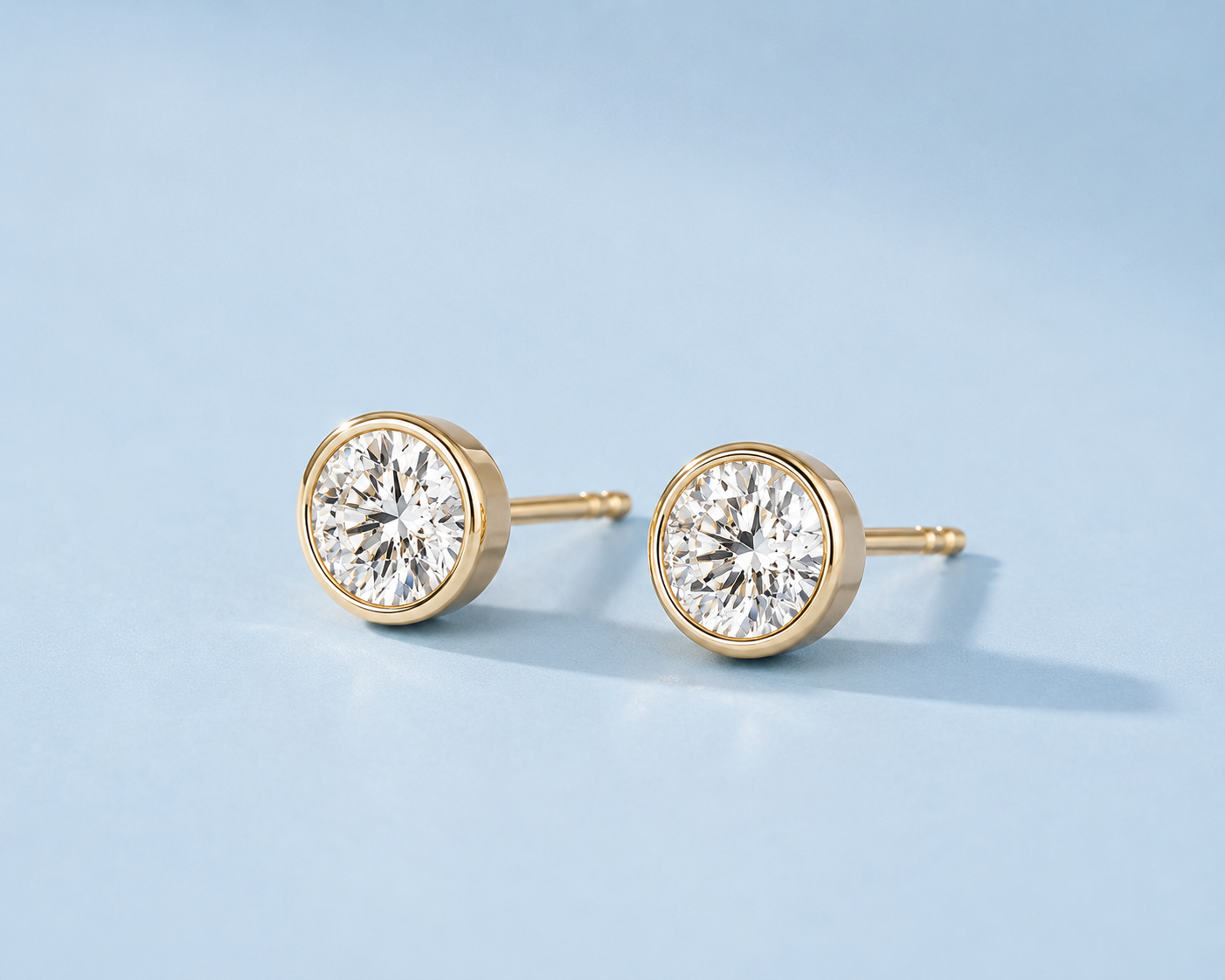 Angled view of gold bezel-set stud earrings with round clear stones on a light blue background