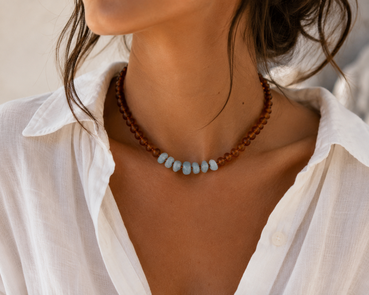 A close-up of a woman wearing the Hollis Necklace with pale blue raw aquamarine chips and warm cognac Baltic amber beads, styled with a white collared shirt.