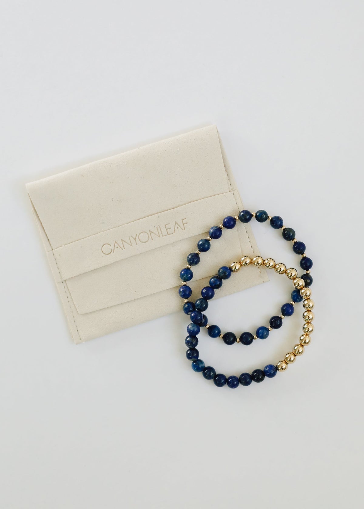 Indi Sun + Moon coordinating lapis lazuli and 14K gold-filled stretch bracelets from The Riann Ashlyn Collection beside a cream jewelry pouch.