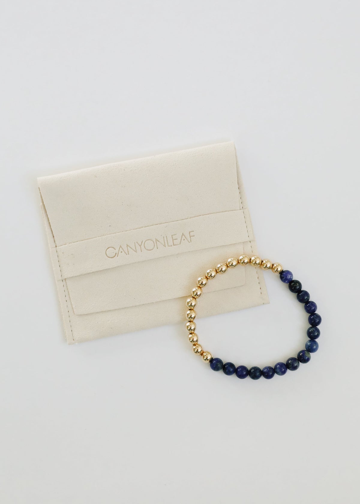 Individual Indi two-tone stretch bracelet from The Riann Ashlyn Collection, featuring 14K gold-filled beads and deep blue lapis lazuli beside a cream jewelry pouch.