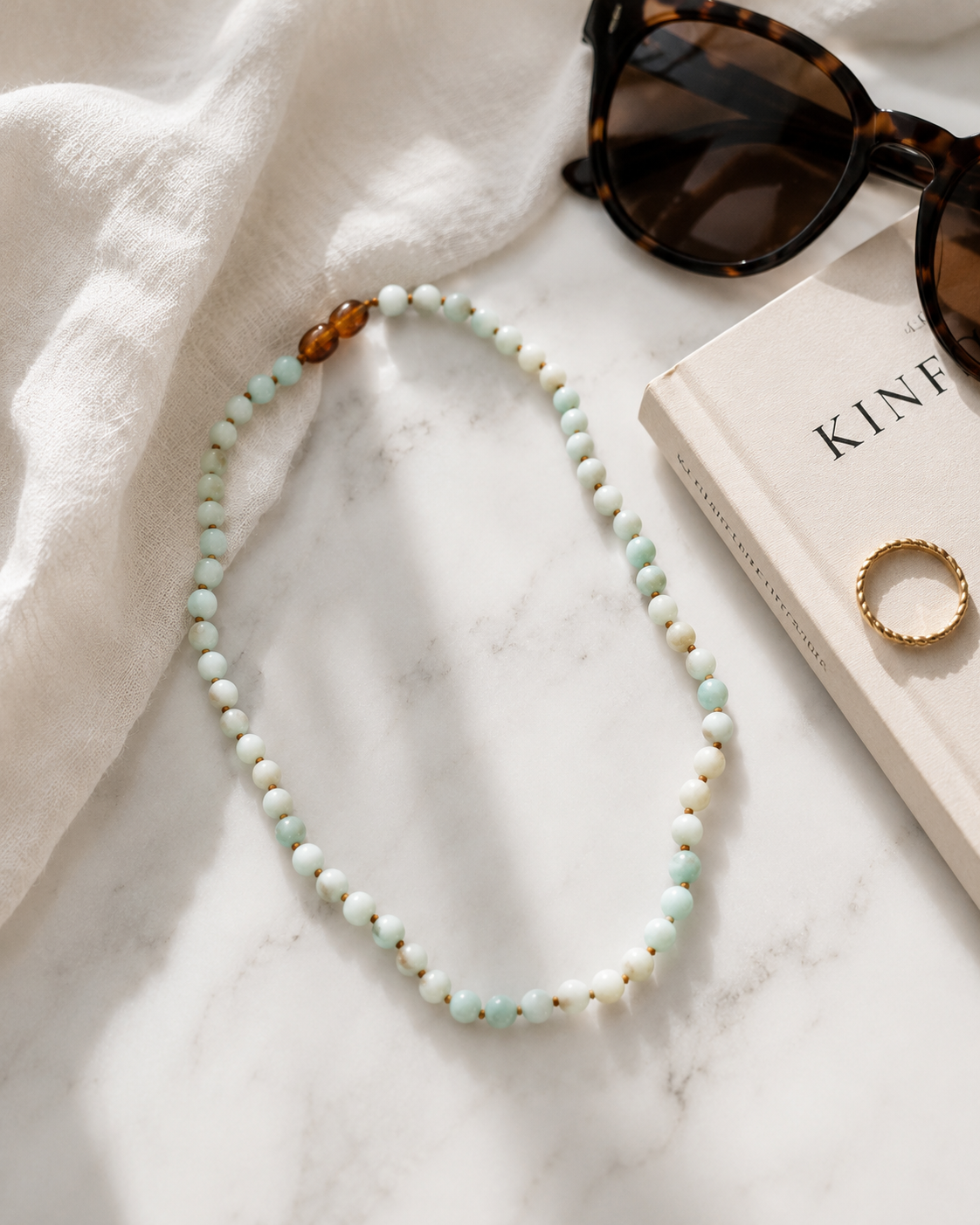 Maren Amazonite Gemstone Necklace from The Riann Ashlyn Collection featuring pale blue-green gemstone beads and amber-brown accents, styled on white marble beside sunglasses, a book, and a gold ring.