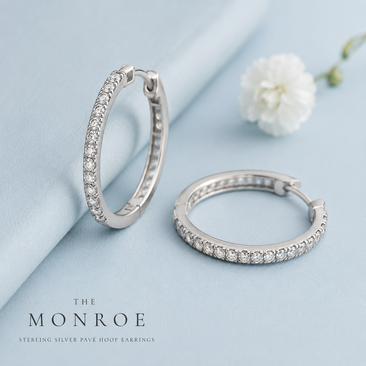 Silver pavé hoop earrings with clear stones displayed on a light blue background with Monroe branding.