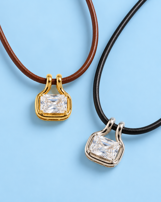 Two Palmer leather cord pendant necklaces on a soft RA blue background, featuring a brown cord with a gold-tone rectangular cubic zirconia pendant and a black cord with a silver-tone rectangular cubic zirconia pendant.
