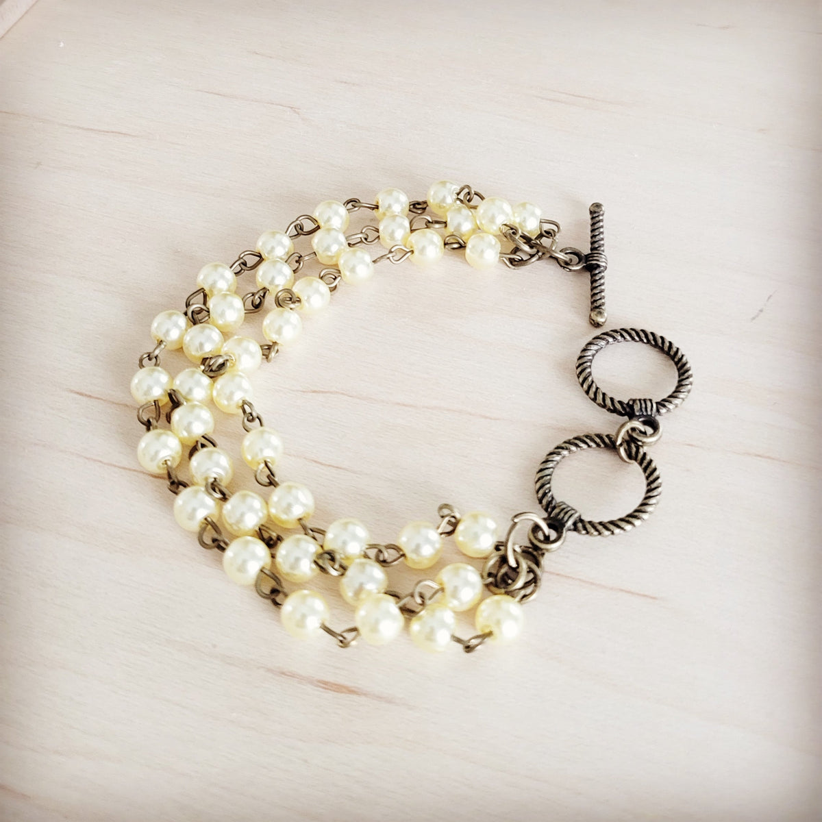 Raye triple strand pearl bracelet with cream glass pearls and antique-tone double-ring toggle clasp on a light wood background