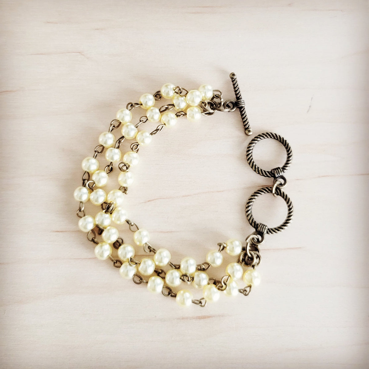 Raye triple strand pearl bracelet with cream glass pearls, antique-tone chain, and double-ring toggle clasp on a light wood background.