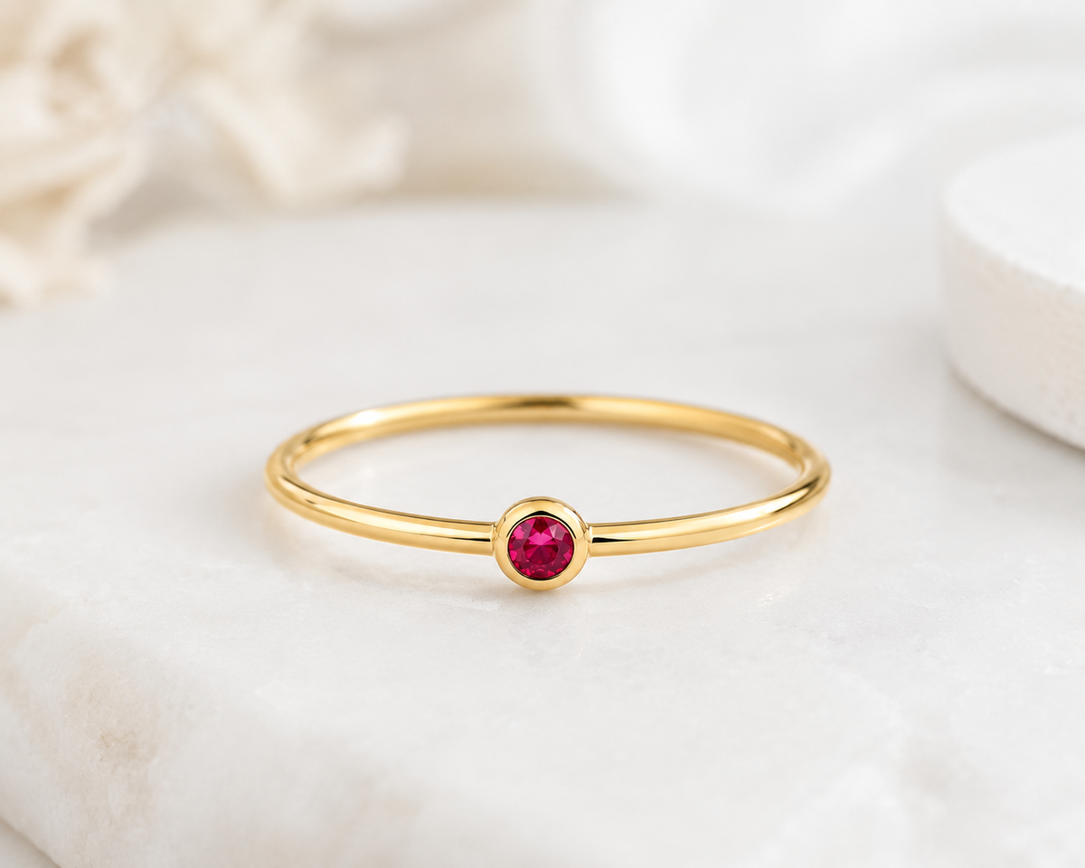     Riann Tiny Ruby Birthstone Ring in yellow gold on a soft neutral background