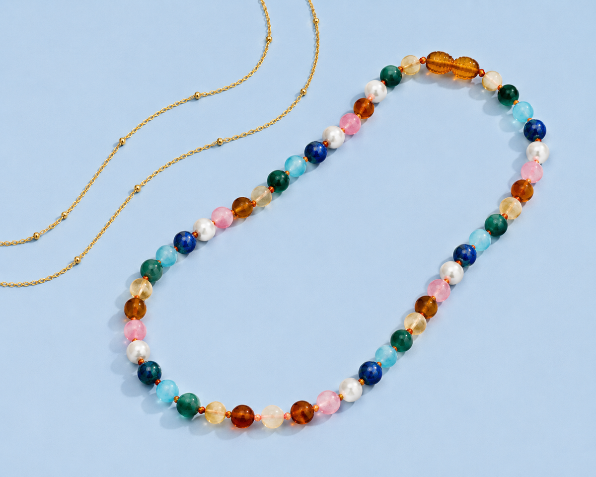 The Sunday Necklace, a colorful beaded necklace featuring Baltic amber, freshwater pearls, and multicolored natural gemstones, displayed on a light blue background beside a delicate gold chain.